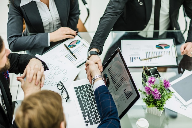 Handschlag von Geschäftspartnern vor dem Hintergrund von am Arbeitsplatz liegenden Finanzgraphen