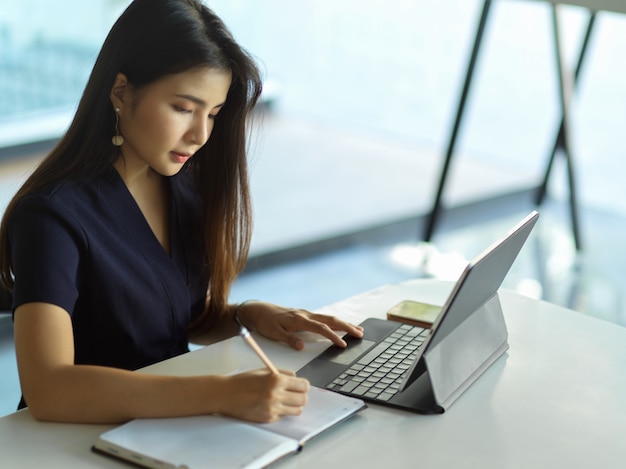 Handschrift der Geschäftsfrau, die auf Notizbuch schreibt, während mit Laptop auf dem Tisch arbeitet