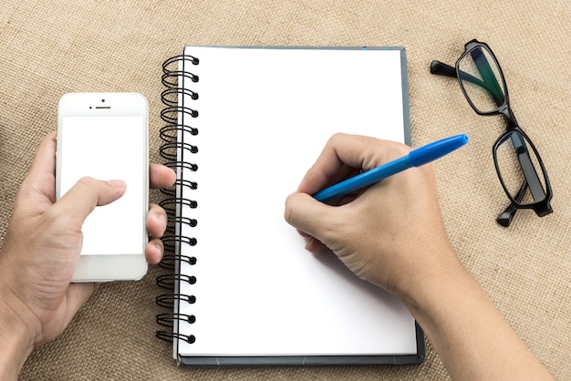 Handschrift des jungen Mannes auf leerem Notizbuch, während eine andere Hand Smartphone auf hölzernem Vorsprung hält