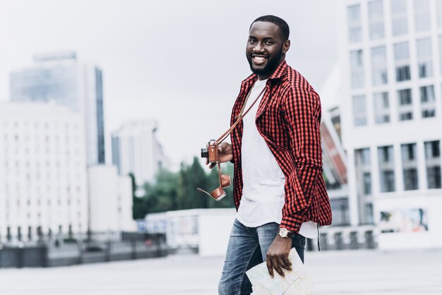 Handsome und glücklich Afro American Tourist mit alten Kamera und Karte in der modernen Stadt