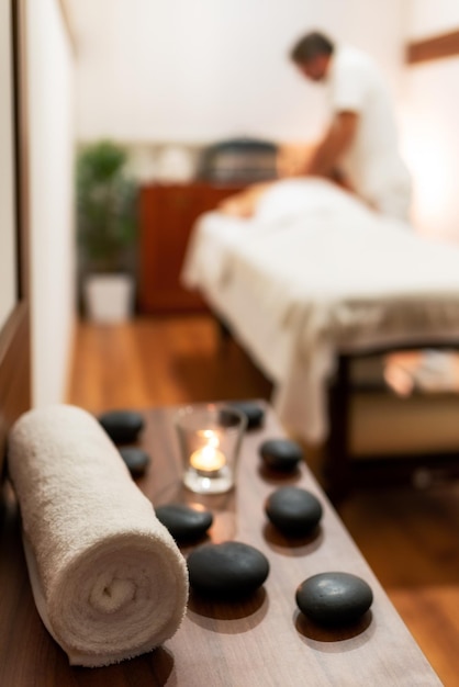 Handtuchkerze und Lavasteine im Spa-Salon Verschwommener Masseur mit Kunde, der im Hintergrund auf dem Massagetisch liegt