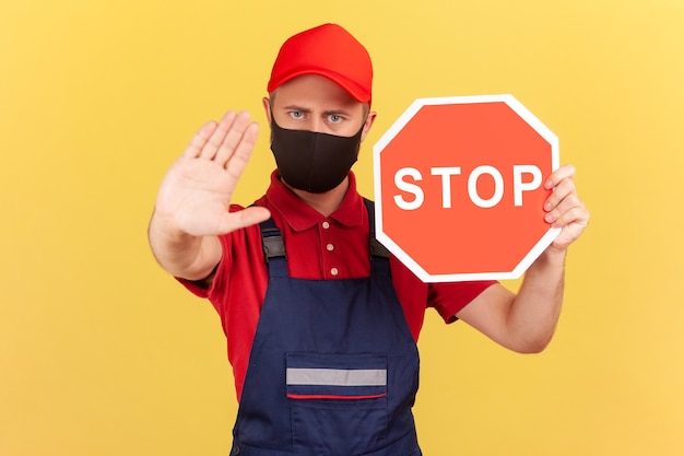 Handwerker in einheitlicher und hygienischer Gesichtsmaske, die Stoppgeste zeigt und rotes Stoppschild hält,