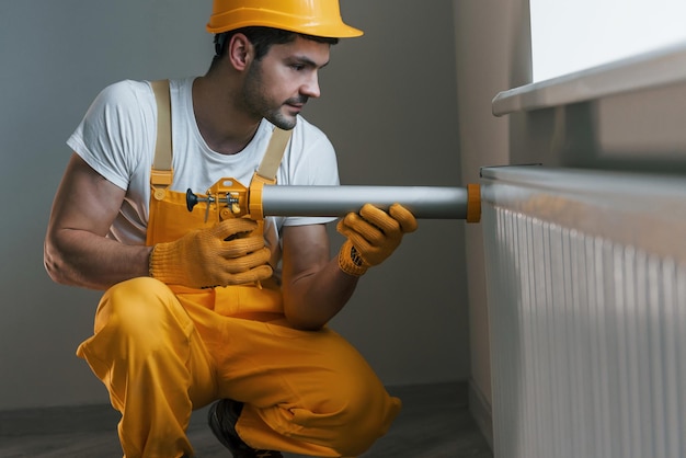 Handwerker in gelber Uniform arbeitet im Innenbereich mit Wärmebatterie, indem er ein spezielles Werkzeug verwendet Hausrenovierungskonzeption