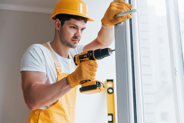 Handwerker in gelber Uniform installiert neues Fenster mit automatischem Schraubendreher Konzeption der Hausrenovierung