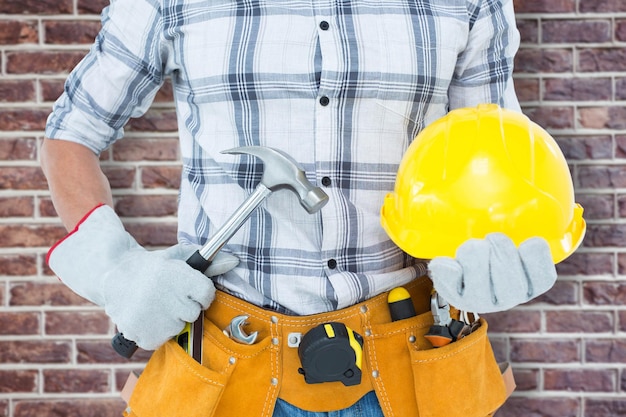 Handyman segurando martelo e capacete contra a parede de tijolos vermelhos