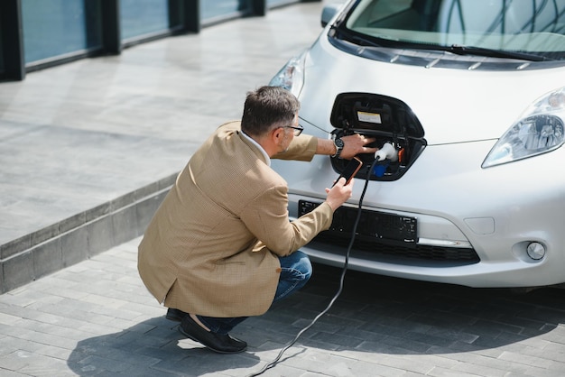 Hansome bärtiger Mann, der neben seinem neuen modernen Elektroauto sitzt und den Stecker des Ladegeräts hält, während das Auto an der Ladestation lädt
