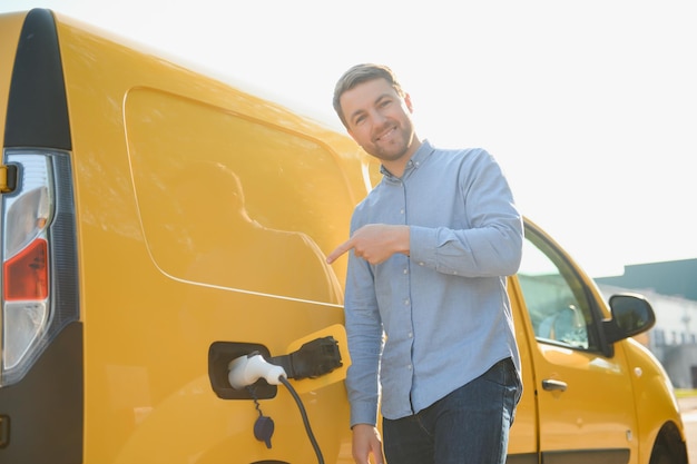 Hansome barbudo perto de seu novo carro elétrico moderno na estação de carregamento