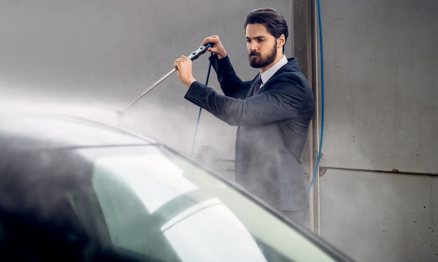 Hansome elegante joven barbudo en traje lavando su automóvil en la estación de autoservicio de lavado manual de automóviles