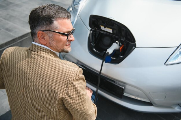 Hansome hombre barbudo sentado cerca de su nuevo coche eléctrico moderno y sujetando el enchufe del cargador mientras el coche se carga en la estación de carga