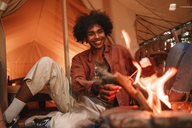 Hansome junger lockiger afrikanischer Mann röstet Marshmallows auf Spießen über einer Feuerstelle auf dem Campingplatz und genießt einen Glamping-Urlaub im Freien mit Freunden, die nach einer Pandemiesperre wiedereröffnet werden.