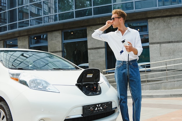 Hansome-Typ sitzt in der Nähe seines neuen modernen Elektroautos und hält den Stecker des Ladegeräts, während das Auto an der Ladestation lädt