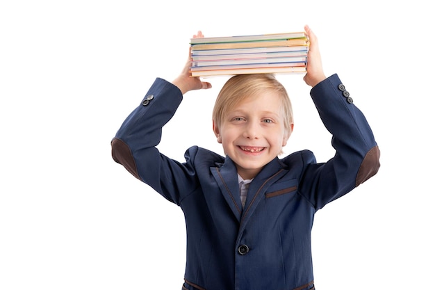 Happy blond Schuljunge in Schuluniform hält Stapel Bücher auf dem Kopf Portrait von Grundschüler isoliert auf weißem Hintergrund