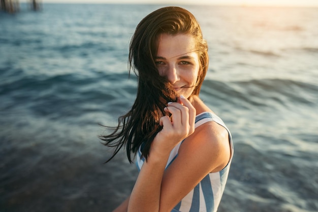 Happy brunette schöne Frau spielen mit Wind weht Haare auf Meer Sonnenuntergang Hintergrund Cover-Idee Sexy Frau im Kleid zu Fuß im Ozean mit gesundem Lächeln Reisekonzept Lifestyle-Mode-Konzept