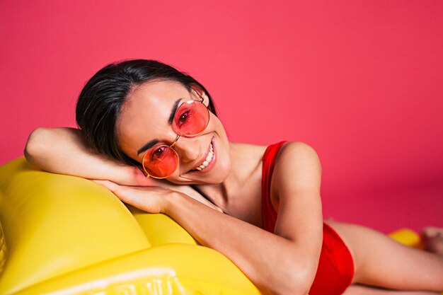 Happy cute brunette Frau liegt auf gelben Luftmatratze in roten Badeanzug und rosa Sonnenbrille isoliert über rosa Hintergrund Sommerzeit und Urlaub Entspannen Sie sich am Strand