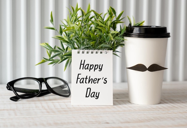 Happy Fathers Day Konzept Kaffeetasse mit Papierherzanhänger mit Happy Father's Day Text und Notebook-Brille auf Holztischhintergrund