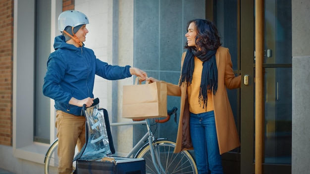 Happy Food Delivery Hombre que lleva una mochila térmica en una bicicleta entrega el pedido de un restaurante a una cliente femenina Correo entrega almuerzo para llevar a una chica hermosa en el edificio de la Oficina del Distrito de la Ciudad Moderna