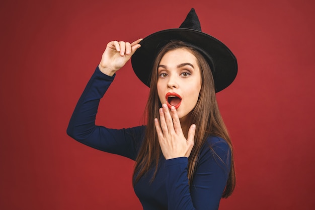 Happy Halloween Witch mit hellem Make-up und langen Haaren. Schöne junge überraschte Frau, die im sexy Kostüm der Hexen aufwirft. Auf rotem Hintergrund isoliert.