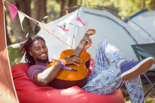 Happy hipster relajante en camping tocando la guitarra