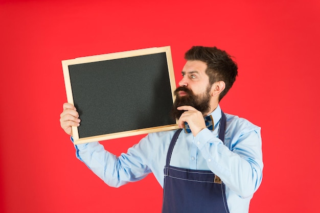 Happy-Hour-Bar. Hipster-Barkeeper zeigen Tafelkopierraum. Personal im Hipster-Restaurant. Hipster informiert dich. Mann bärtiger Barkeeper oder Koch in Schürze halten leere Tafel. Konzept der Preisliste.