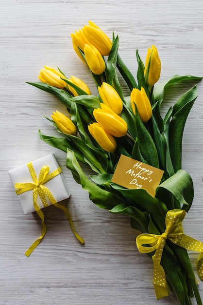 Happy Mother's Day Textschild auf Bastelpostkarte Tulpenstrauß mit Geschenkbox auf weißem Holzhintergrund Feiertagsgrußkarte zum Muttertag Draufsicht flach gelegt