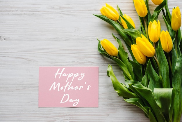 Happy Mother's Day Textschild mit Bouquet gelbe Tulpen auf weißem rustikalem Holzhintergrund Grußkarte mit Blumenkonzept Urlaubsgrußkarte zum Muttertag Draufsicht flach gelegt