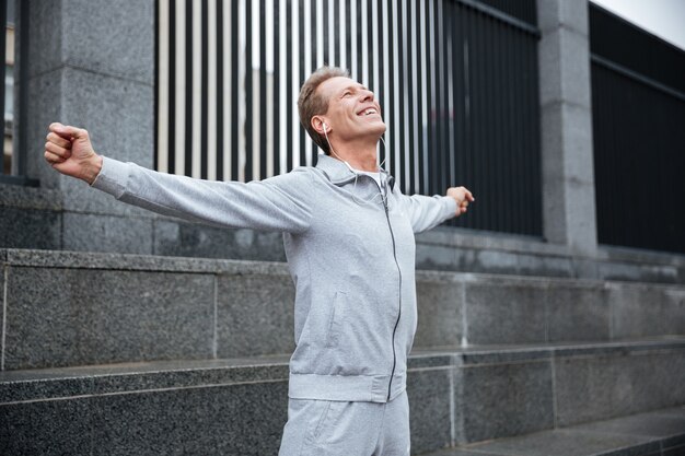 Happy Runner en ropa deportiva gris y auriculares calentándose en la calle con las manos a los lados.