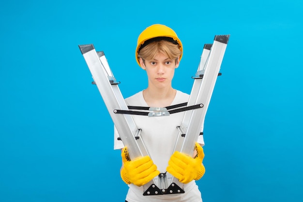 Happy Teen in weißen T-Shirt und Handschuhe mit gelben Helm im Kopf Leiter in den Händen halten stehend im Studio auf blauem Hintergrund Ein zukünftiger Architekt isoliert auf blauem Hintergrund