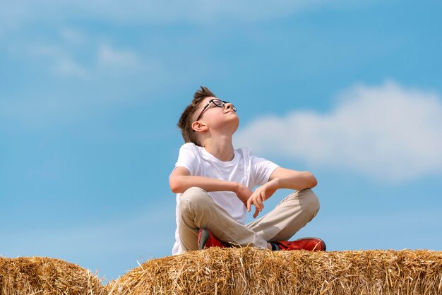 Happy Teenager sitzt auf Heuhaufen gegen den blauen Himmel Kind genießt die frische Luft und die Sonne im Dorf