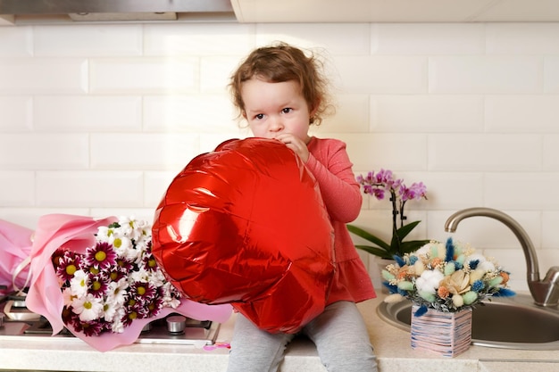 Happy Valentine's Day Süßes Mädchen mit rotem Herzen, das zu Hause auf dem Küchentisch mit Blumen sitzt