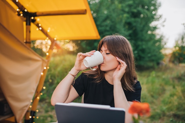 Happy Woman Freelancer mit Laptop und Kaffeetrinken in einem gemütlichen Glamping-Zelt an einem sonnigen Tag Luxus-Campingzelt für Sommerferien und Urlaub im Freien Lifestyle-Konzept