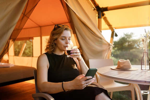 Happy Woman Freelancer nutzen Telefon und trinken Cocktailanrufe, während sie sich an sonnigen Tagen in einem gemütlichen Glamping-Zelt entspannen Luxus-Campingzelt für Sommerferien und Urlaub im Freien Lifestyle-Konzept