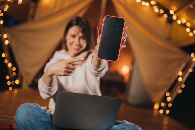 Happy Woman Freelancer zeigt Telefon, während sie in der Sommernacht auf einem gemütlichen Glamping-Zelt mit Laptop arbeitet Luxus-Campingzelt für Outdoor-Urlaub und Urlaub Lifestyle-Konzept