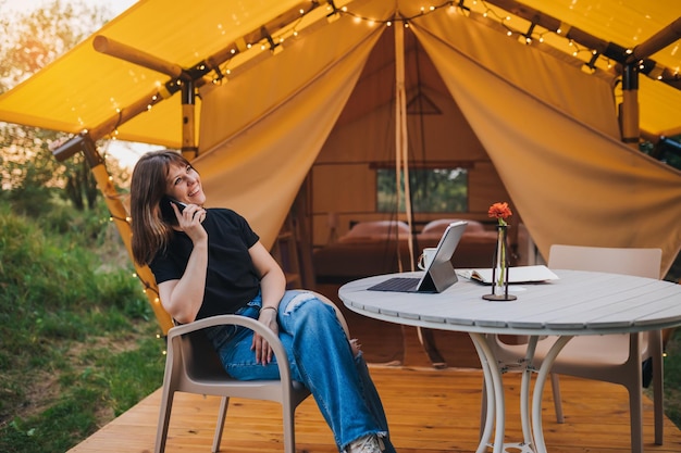 Happy Woman Freiberufler sprechendes Telefon und mit Laptop auf gemütlichem Glamping-Zelt an einem sonnigen Tag Luxus-Campingzelt für Sommerferien und Urlaub im Freien Lifestyle-Konzept