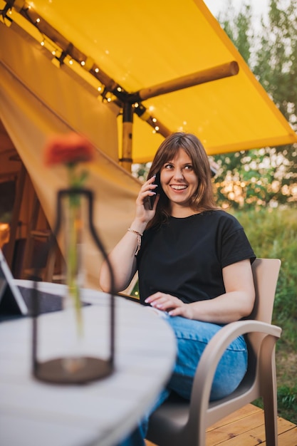 Happy Woman Freiberufler sprechendes Telefon und mit Laptop auf gemütlichem Glamping-Zelt an einem sonnigen Tag Luxus-Campingzelt für Sommerferien und Urlaub im Freien Lifestyle-Konzept