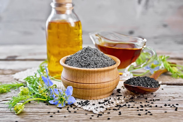 Harina de alcaravea negra en un cuenco, semillas en una cuchara, aceite en una botella y salsera sobre arpillera, ramitas de Nigella sativa con flores azules y hojas sobre tabla de madera