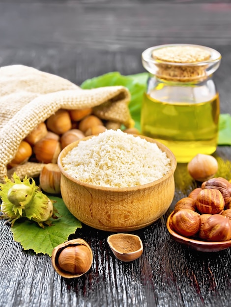 Harina de avellana en un cuenco, nueces en bolsa, una cuchara, aceite en frasco de vidrio y rama de avellana con hojas verdes sobre fondo de tablero de madera negro