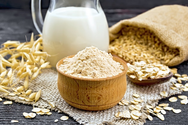 Harina de avena en un cuenco, leche en una jarra, avena en una cuchara sobre arpillera, grano en bolsa, tallos de avena sobre fondo de tabla de madera