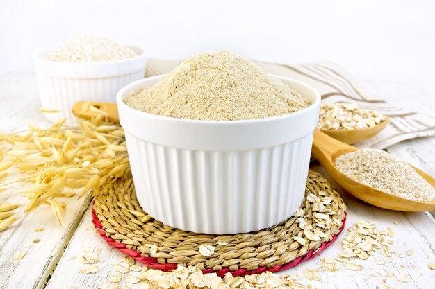 Harina Oaten en tazón de fuente blanco, avena y salvado en una cuchara, tallos de avena, servilleta con el telón de fondo de tablas de madera