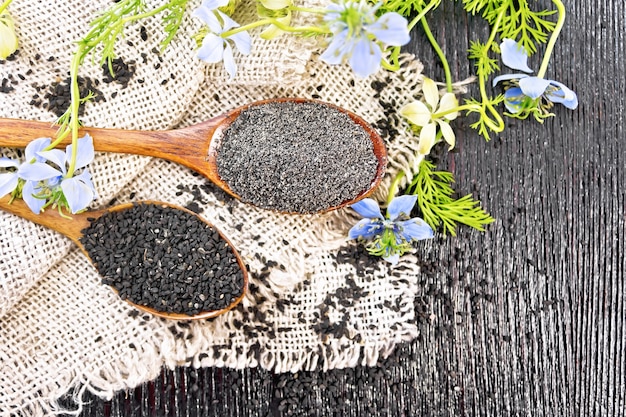 Harina y semillas de Nigella sativa en dos cucharas de arpillera, ramitas de kalingini con flores azules y hojas verdes sobre fondo de una vieja tabla de madera desde arriba