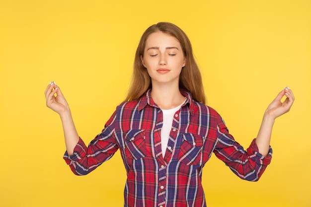 Harmonia e paz interior. Retrato de menina ruiva feliz e calma em camisa quadriculada praticando ioga, de mãos dadas em gesto de namastê, meditando com os olhos fechados. tiro de estúdio isolado em fundo amarelo