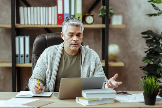 Hart arbeitender Geschäftsmann. Ernsthafter netter Mann, der vor dem Laptop sitzt, während er Notizen im Notizbuch macht