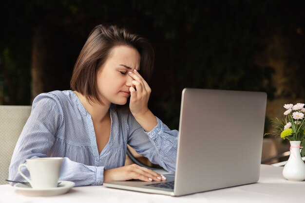 Harte Arbeit zu Hause, Kopfschmerzen. Unglückliche Frau hält Kopf während der Arbeit an einem Laptop drinnen