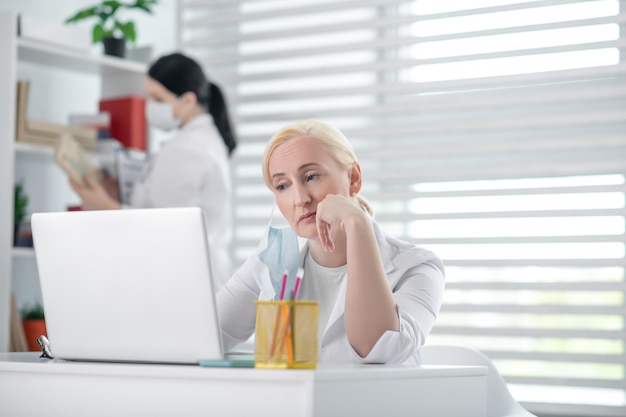 Harter Tag. Müde blonde Ärztin sitzt an einem Laptop, dunkelhaarige Kollegin in einer Schutzmaske in der Nähe des Regals.