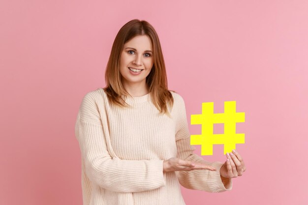 Hashtag y blogs. Retrato de una atractiva mujer rubia feliz sosteniendo un gran símbolo de hash de papel y sonriendo a la cámara, usando un suéter blanco. Disparo de estudio interior aislado sobre fondo rosa.