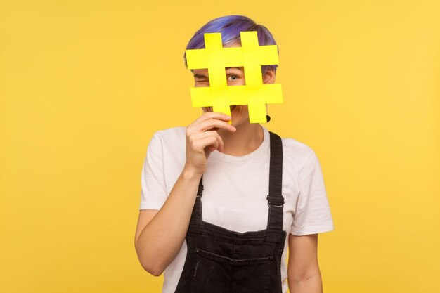 Hashtag, tendencias mediáticas interesantes. Retrato de una joven alegre con el pelo corto violeta en overoles de mezclilla mirando la cámara a través de un gran cartel de hash con ojos curiosos. fondo amarillo, foto de estudio