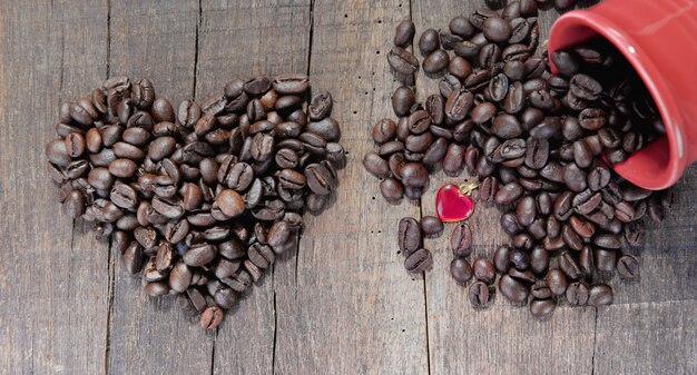 Haufen Kaffeebohnen, die ein Herz und eine Tasse bilden, verschüttet mit einem Juwelenherz, das auf einem rustikalen Brett geformt wird