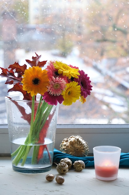 Haufen rosa magentafarbener Koralle und gelber Gerbera-Blumen auf einem Fensterbrett Vergoldete metallische Goldblätter und Kürbisse mit Textilschal auf cremefarbener Fensterbank