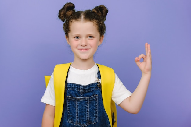 Hausaufgaben sind einfach! Porträt eines süßen süßen Schulmädchens, das ein ok Handzeichen zeigt, einen gelben Rucksack trägt, glücklich in die Kamera schaut und einzeln auf violettem Hintergrund posiert. Kind empfiehlt beste Bildungswahl