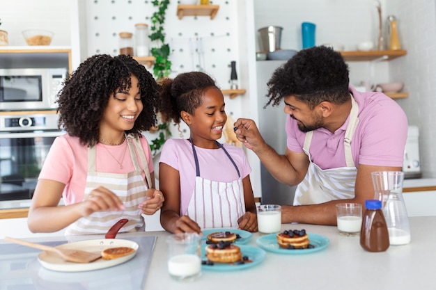 Hausbäckerei zubereiten Liebevolle, freundliche afroamerikanische Familienehepartner mit Tochter, die gemeinsam am Küchentisch Essen kochen und hausgemachte Pfannkuchen backen