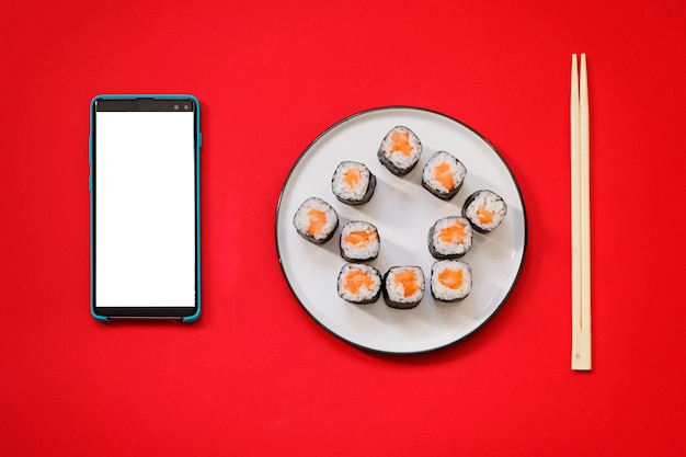 Hausform-Sushi-Rollen mit Stäbchen und Smartphone auf rotem Hintergrund